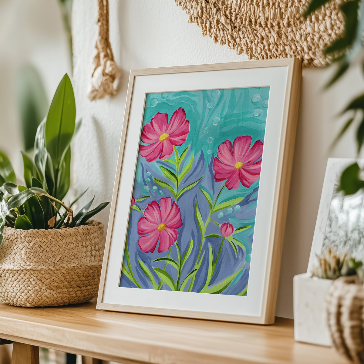 A framed floral art print leaning on a wooden table in a styled home setting, surrounded by potted plants and woven decor. The print features three bright pink flowers with green stems on a textured teal and blue background, enclosed in a light wood frame with a white mat.
