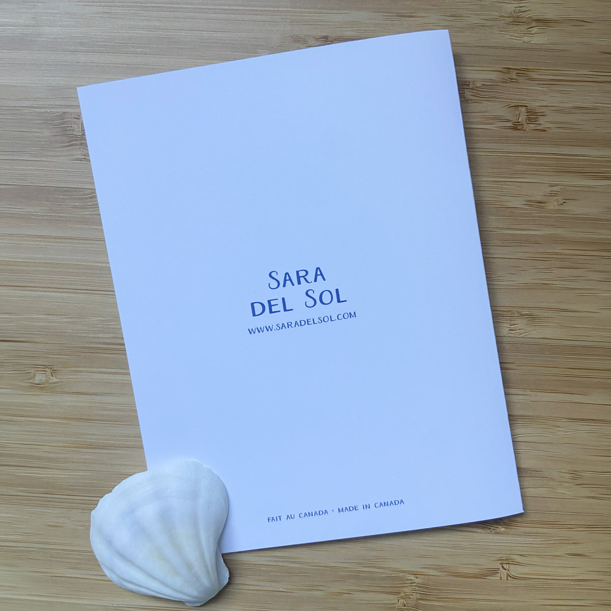 A greeting card background with the brand name 'SARA DEL SOL' and website in blue, placed on a light wood surface with a white seashell in the corner.