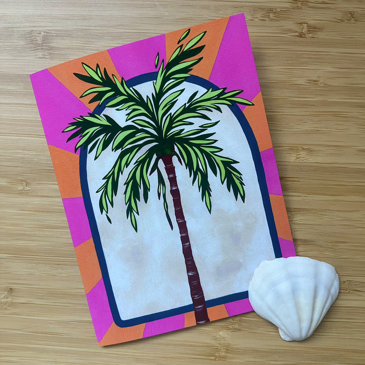 A greeting card featuring a stylized green palm tree, set against a colorful geometric background of pink and orange displayed on a light wood surface next to a white seashell.
