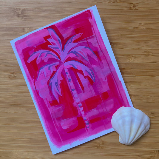 A greeting card featuring a stylized pink and purple palm tree on a bright magenta background, placed on a light wood surface next to a white seashell.