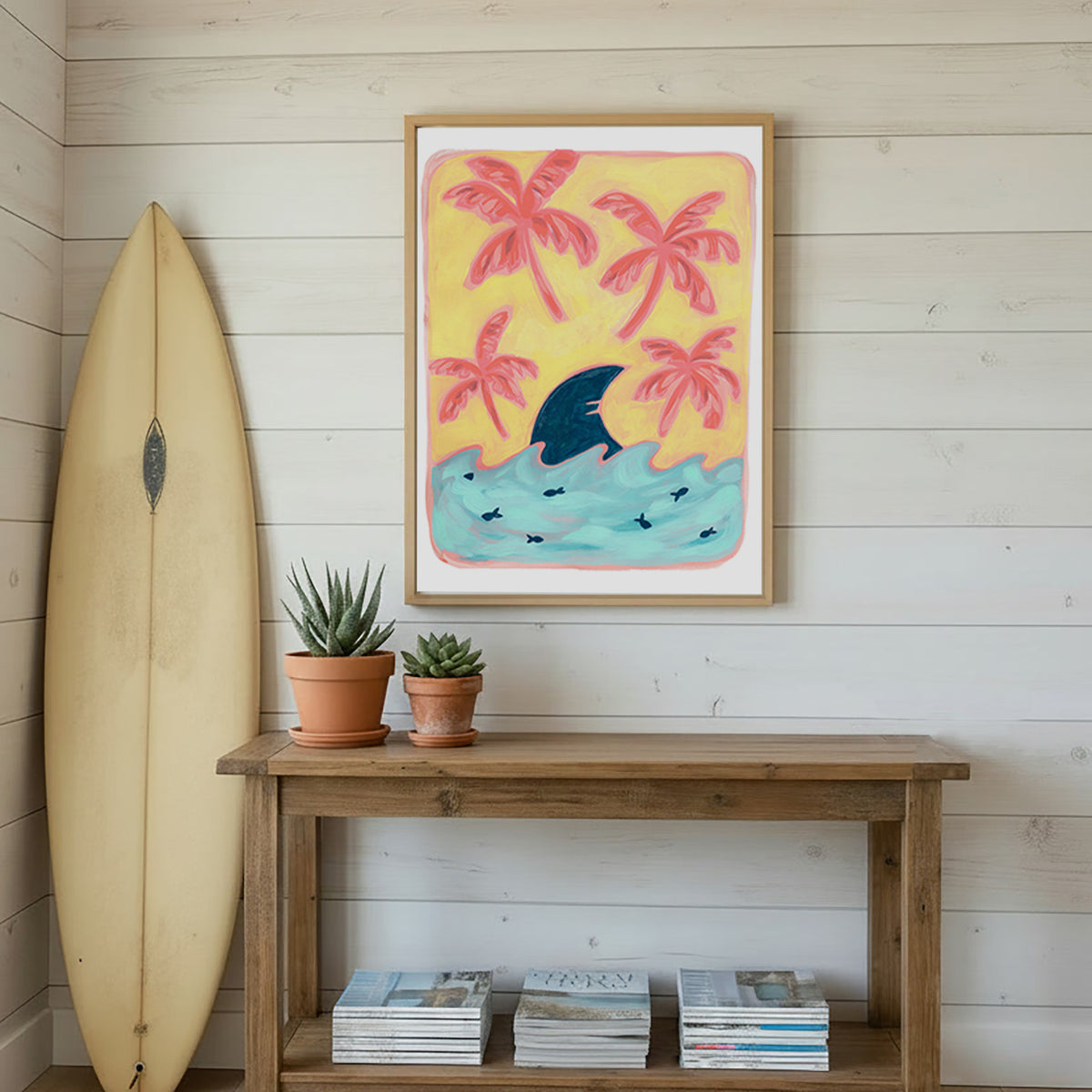 A framed art print featuring a yellow beach scene with pink palm trees, a blue shark fin, and turquoise waves, displayed on a white wood plank wall above a rustic wooden console table with a surfboard and potted succulents.