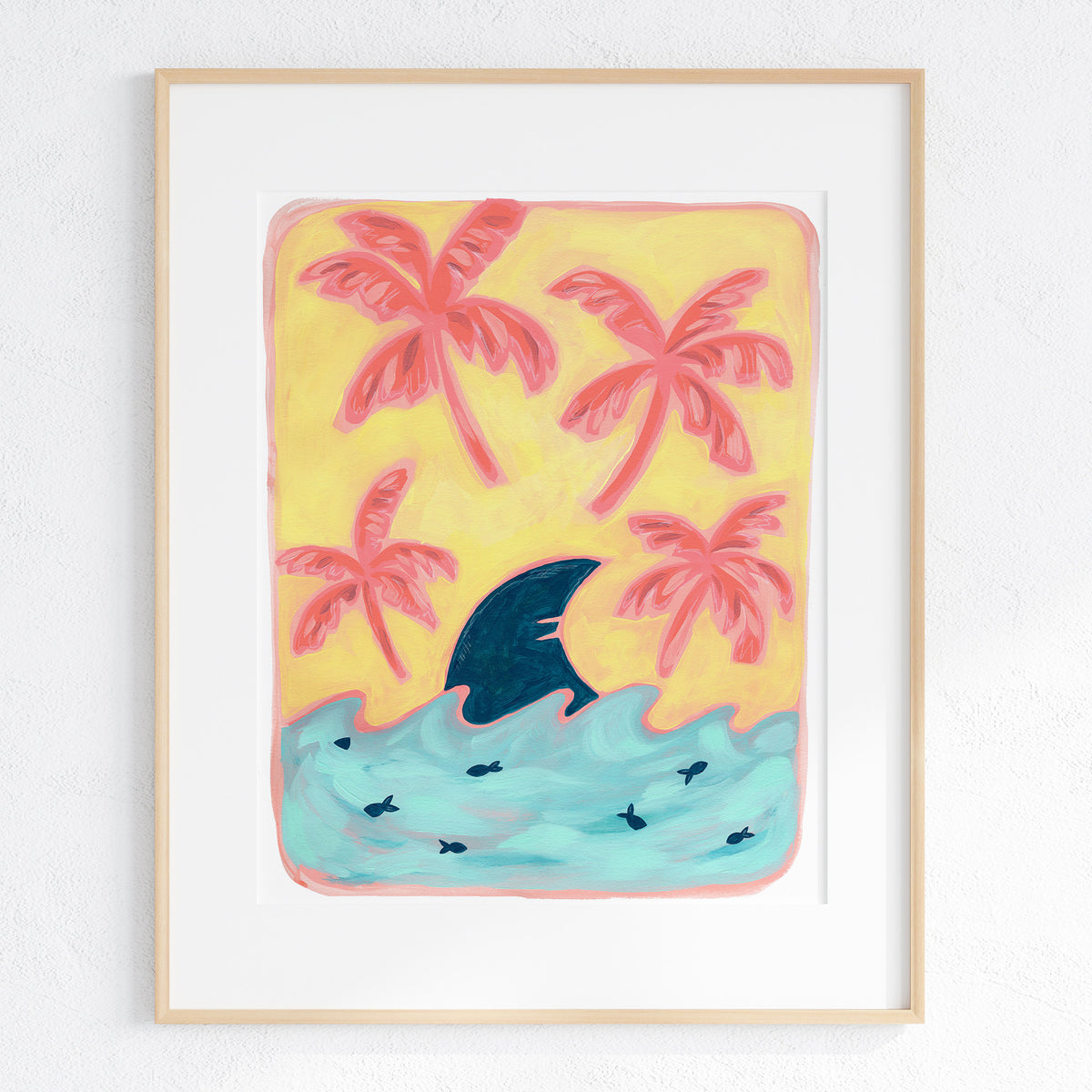 A framed art print featuring a whimsical illustration of pink palm trees, a yellow sky, and turquoise water with a dark blue shark fin breaking the surface, displayed on a textured white wall.
