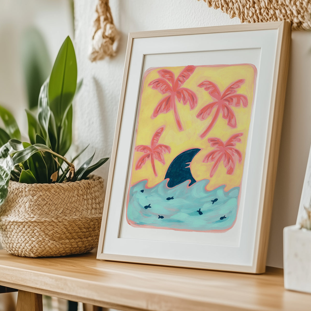 A framed art print leaning on a wooden table next to a potted plant. The print features a yellow background with pink palm trees, a blue shark fin, and turquoise water.