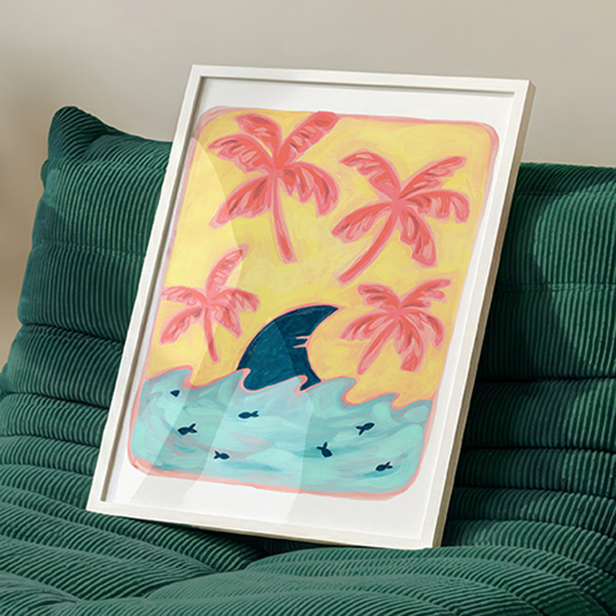 A framed art print featuring a whimsical tropical scene with pink palm trees, a dark blue shark fin, and light blue waves on a yellow background, styled leaning against a green corduroy sofa.