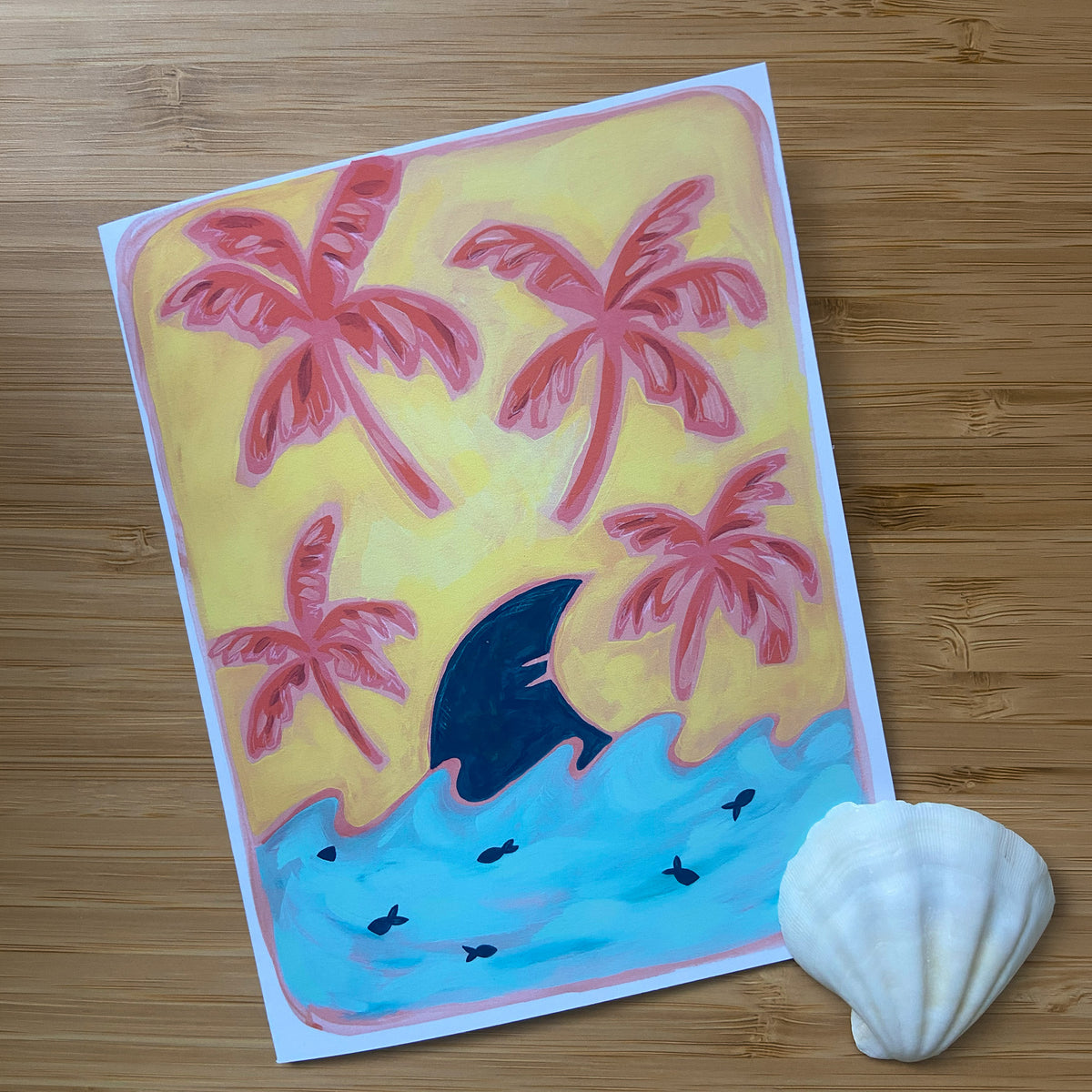 A colorful greeting card depicting a tropical beach scene with pink palm trees, a dark blue shark fin, and light blue water, displayed on a wooden surface next to a white seashell.