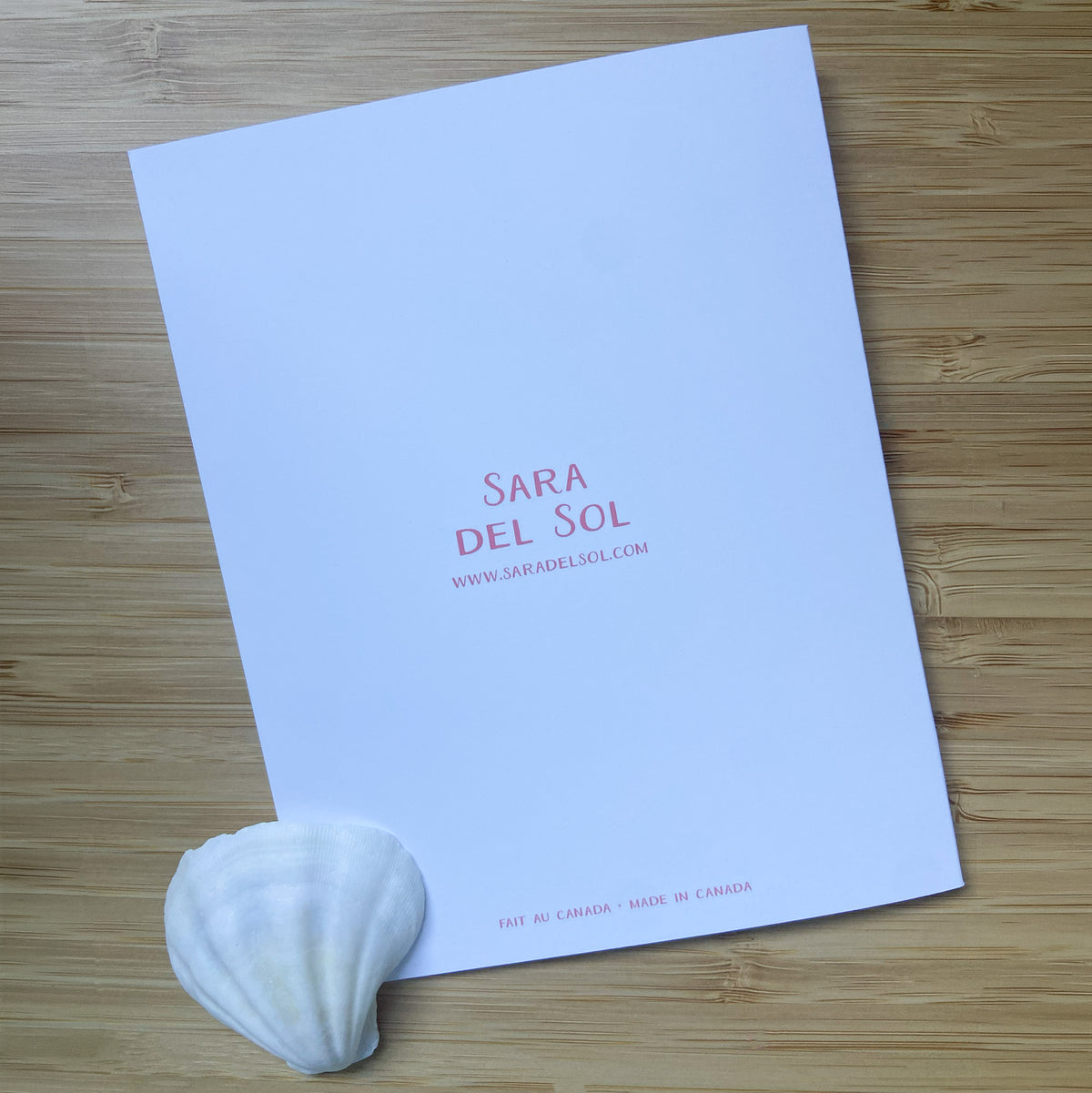 The back of a greeting card with text reading 'SARA DEL SOL' and a website, placed on a light wood surface with a white seashell in the lower left corner.