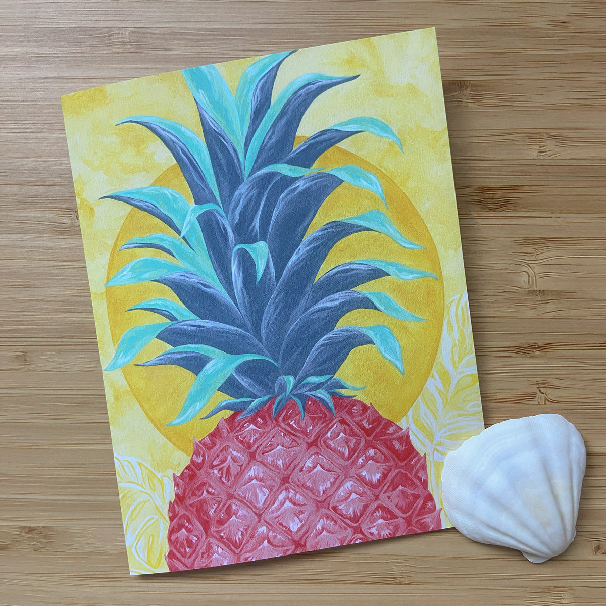 A greeting card featuring a stylized pineapple with a blue and teal crown, pink-red textured body, and a yellow circular background, placed on a light wood surface next to a white seashell.