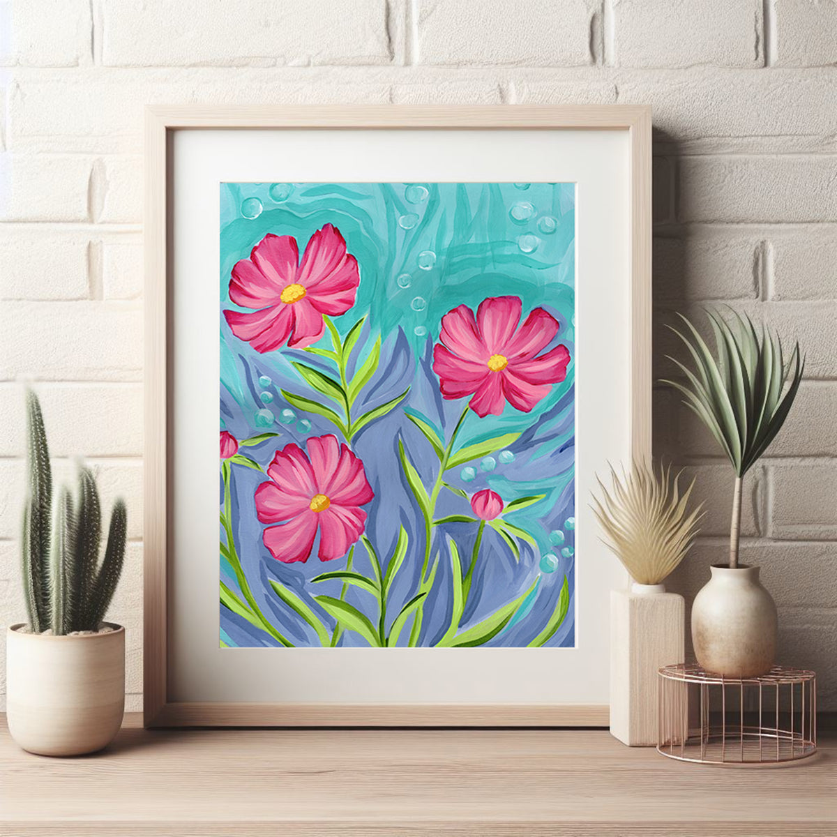 A framed art print featuring three pink flowers with green stems on a turquoise and blue background, styled on a wooden surface against a white brick wall with potted plants.
