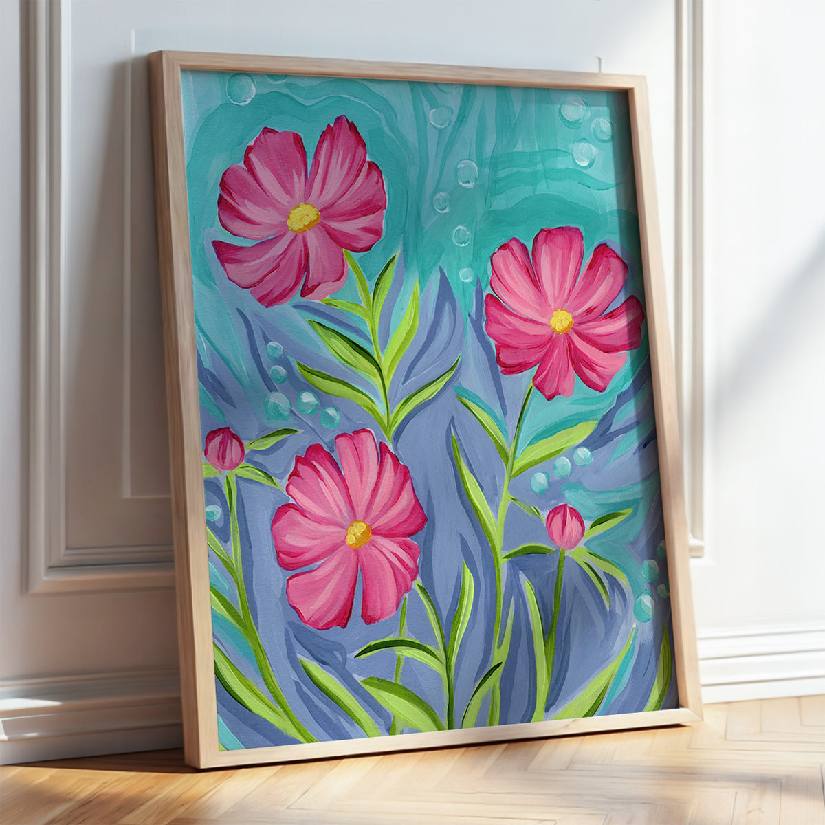 A framed floral painting leaning against a white wall on a light wood floor. The artwork shows three large pink flowers with yellow centers and green stems on a turquoise and blue abstract background.