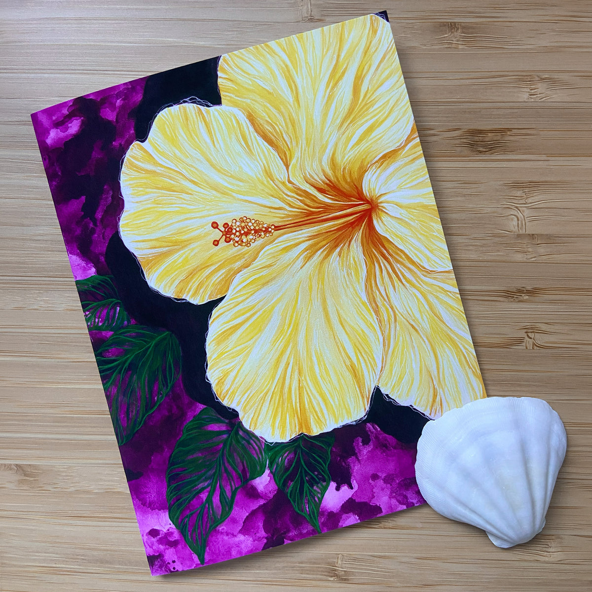 A greeting card featuring a large yellow hibiscus flower with green leaves and a purple watercolor background, displayed on a light wood surface next to a white seashell.
